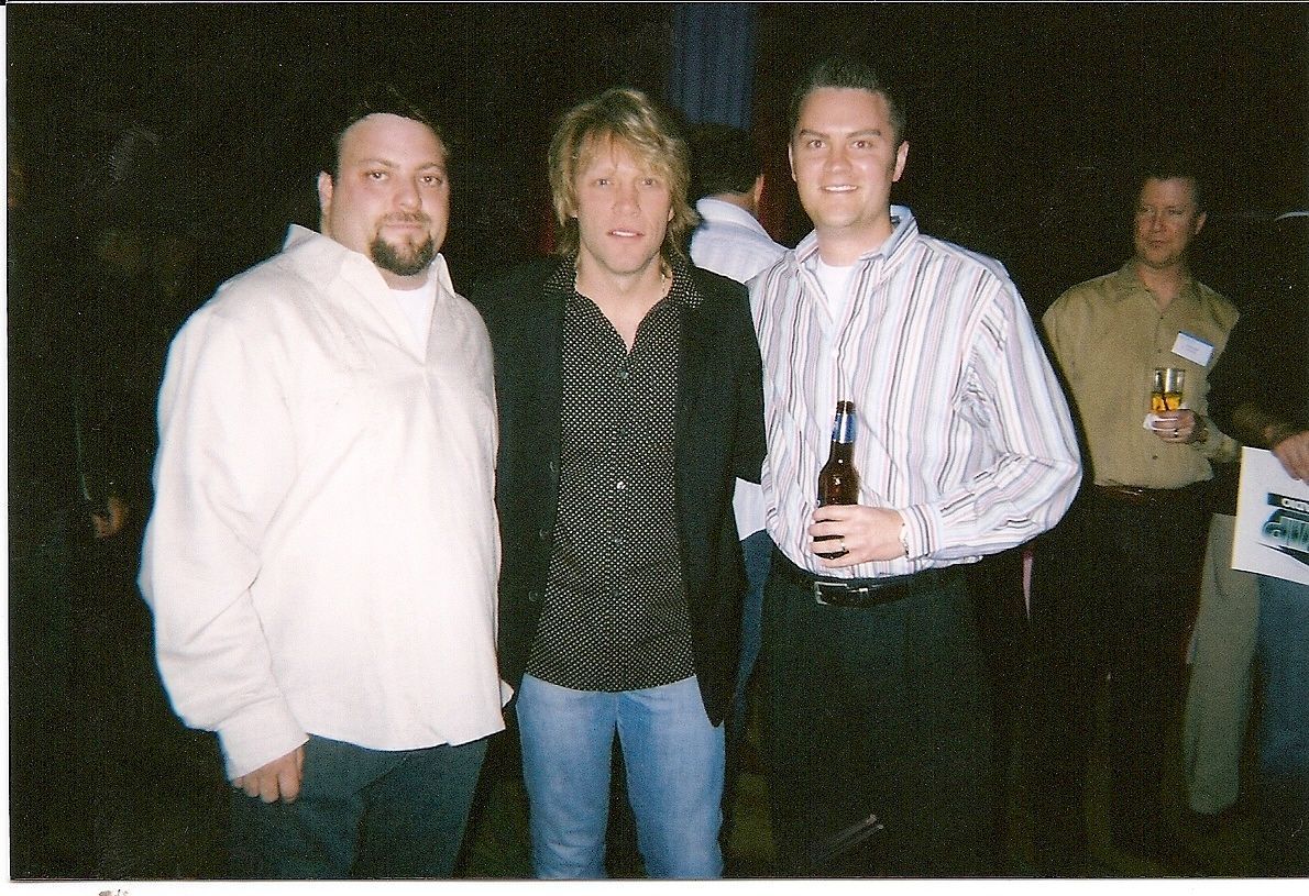 Three men stand together at an indoor event, one holding a beer bottle.