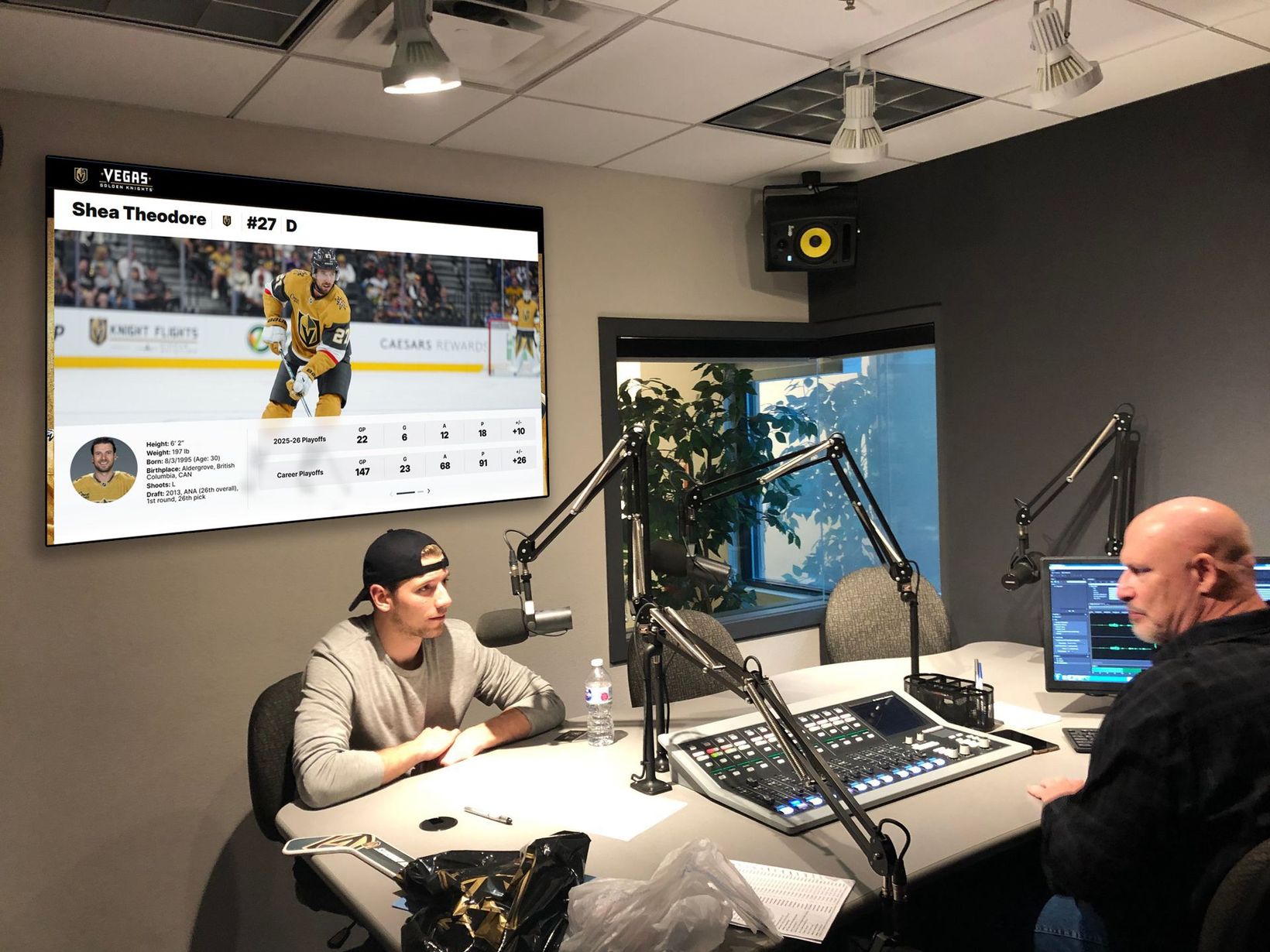 Two people in a radio studio with microphones, mixing board, and a sports game on a wall monitor
