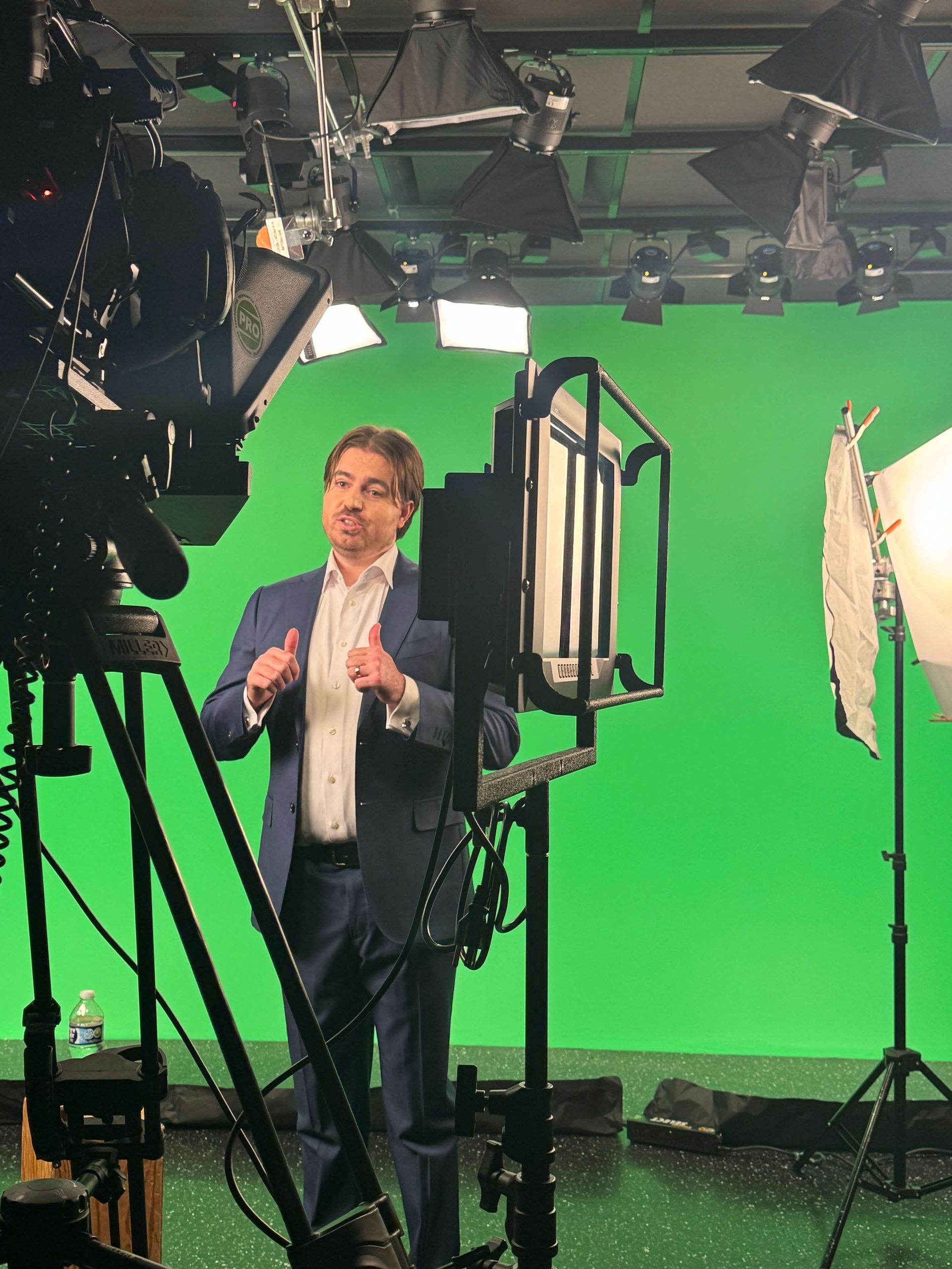 Person speaking in a studio with green screen, cameras, and lighting equipment.