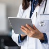 Doctor in white coat holds a tablet, stethoscope visible, indoors.