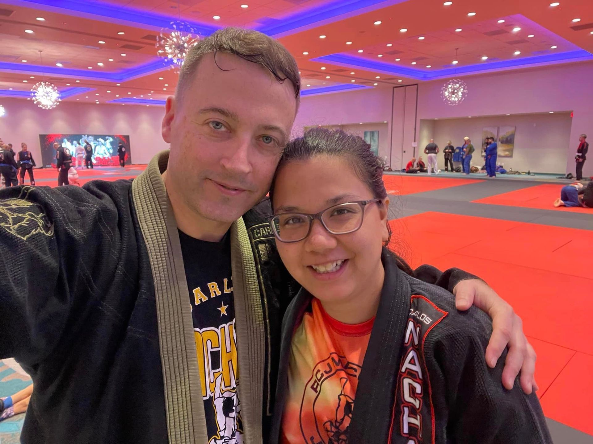 Two people smiling for a selfie at a red-carpet event in a brightly lit ballroom