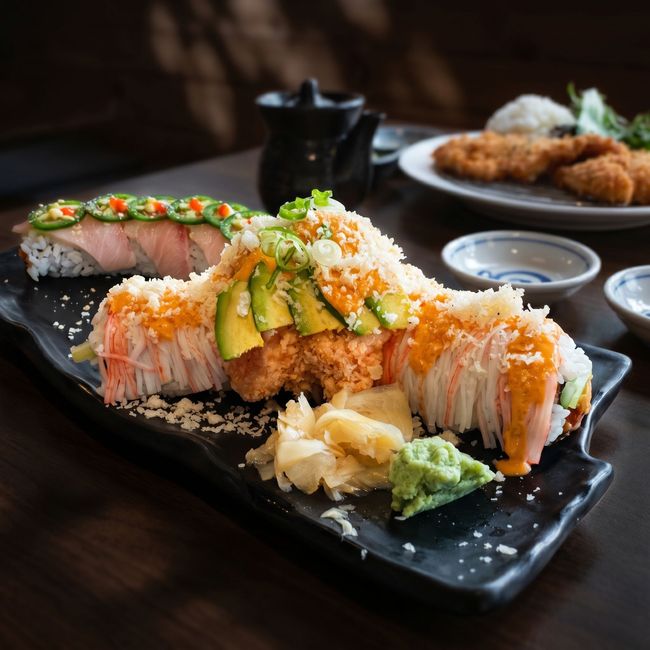 Sushi rolls topped with avocado and crispy flakes on a black plate, with dipping sauce and ginger.