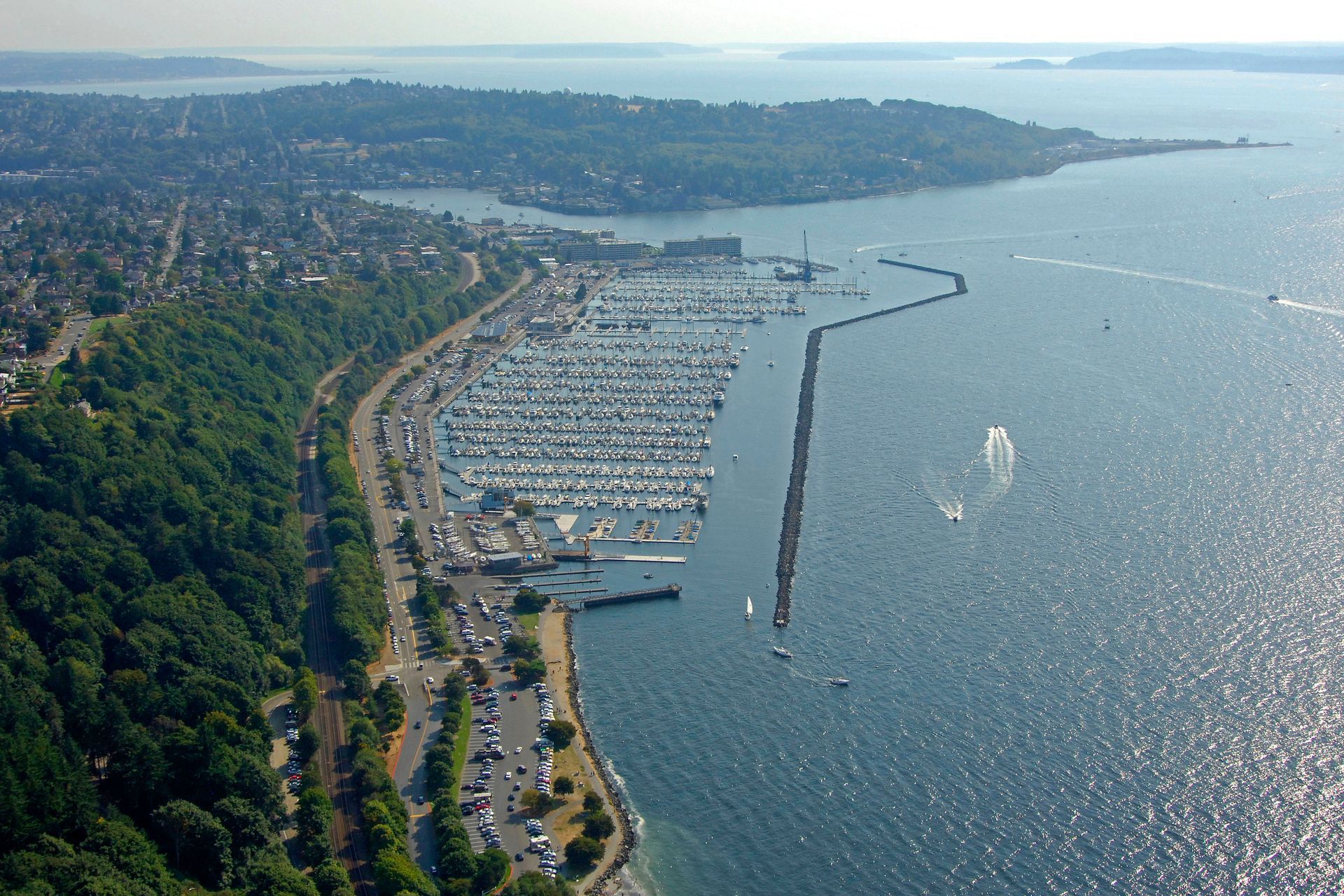 Aerial view of marina filled with boats, next to a tree-lined road and water.