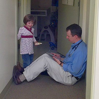 Doctor sitting, reviewing tablet. Child approaches, holding snack in hallway.