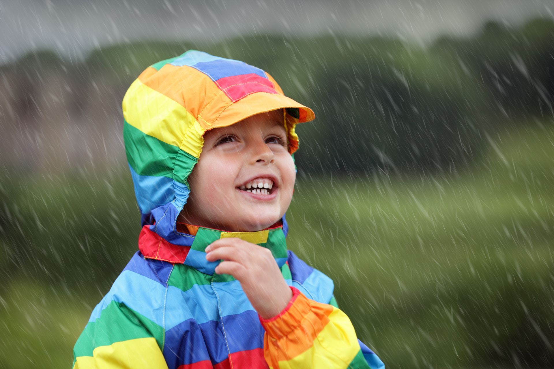 Child in colorful rainbow jacket smiles and looks up at falling rain.