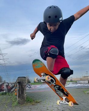 Boy in helmet, knee pads, and red shorts skateboarding at a skatepark, mid-air.