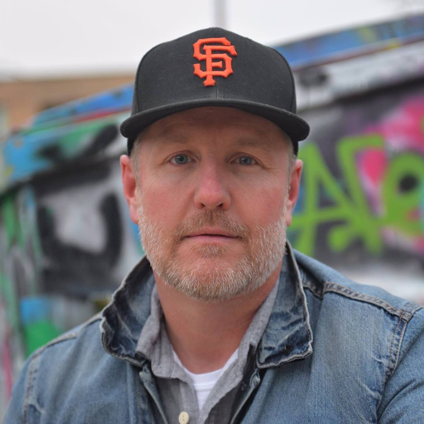 Man in a black SF cap and denim jacket in front of a colorful graffiti wall