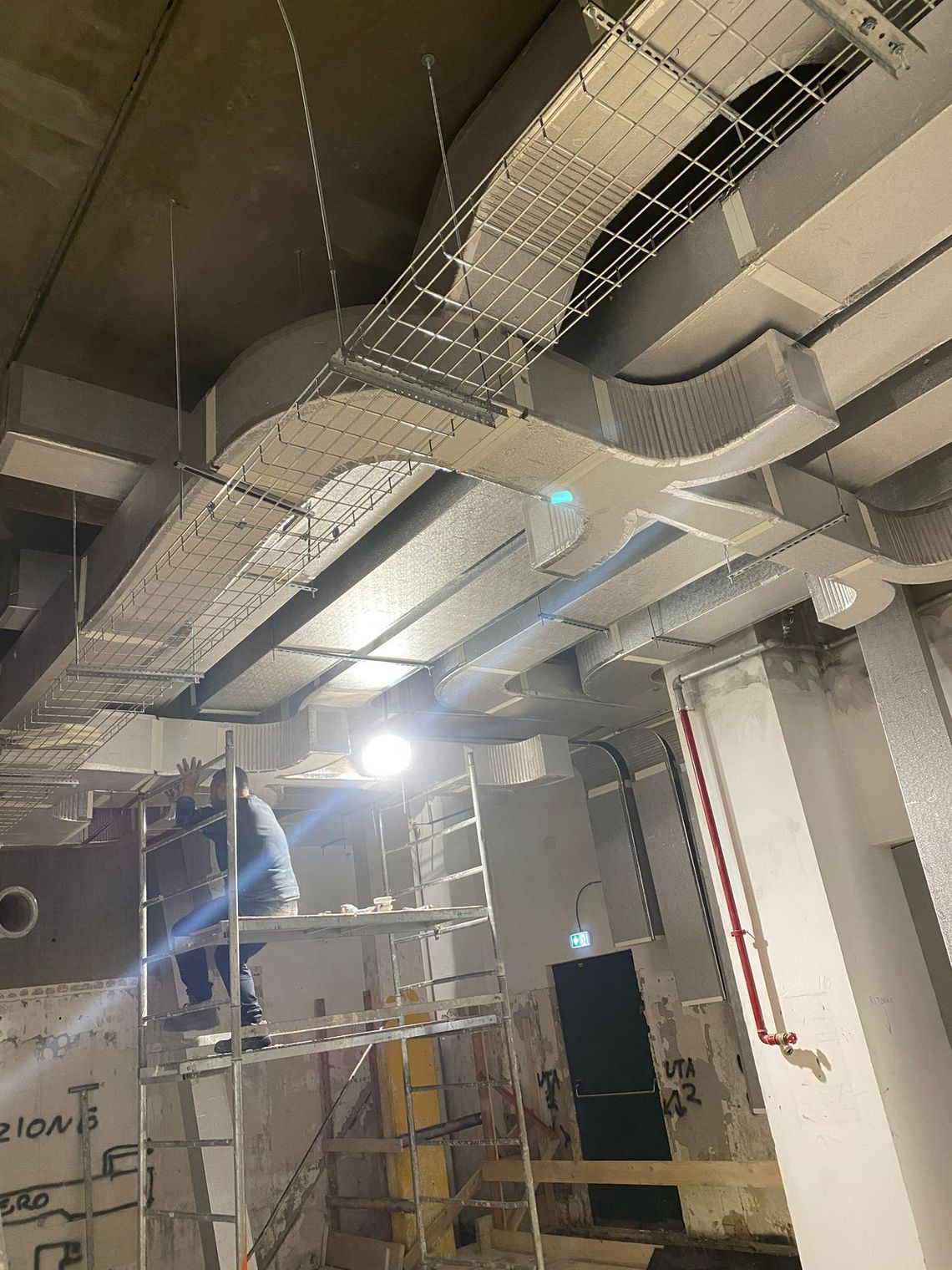 Soffitto di un locale industriale con condotti a vista, canaline portacavi, impalcature e una luce di lavoro intensa.