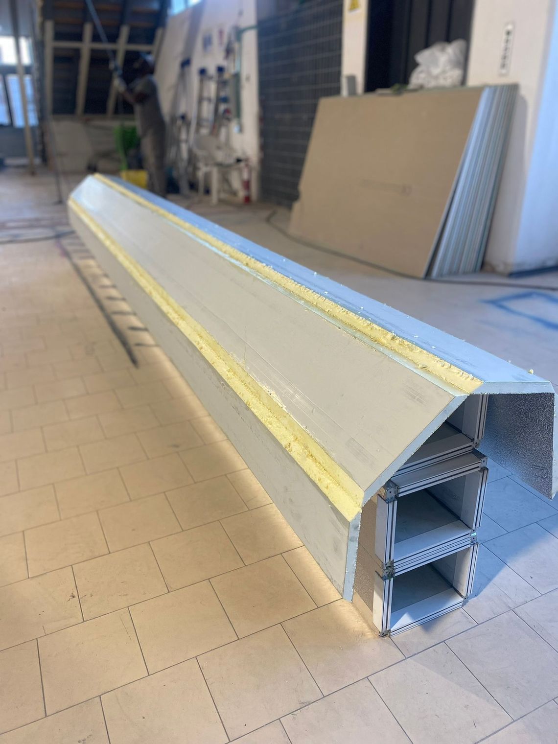 Lungo pannello metallico isolante sul pavimento di un'officina, con nastro adesivo giallo e estremità in schiuma a vista.