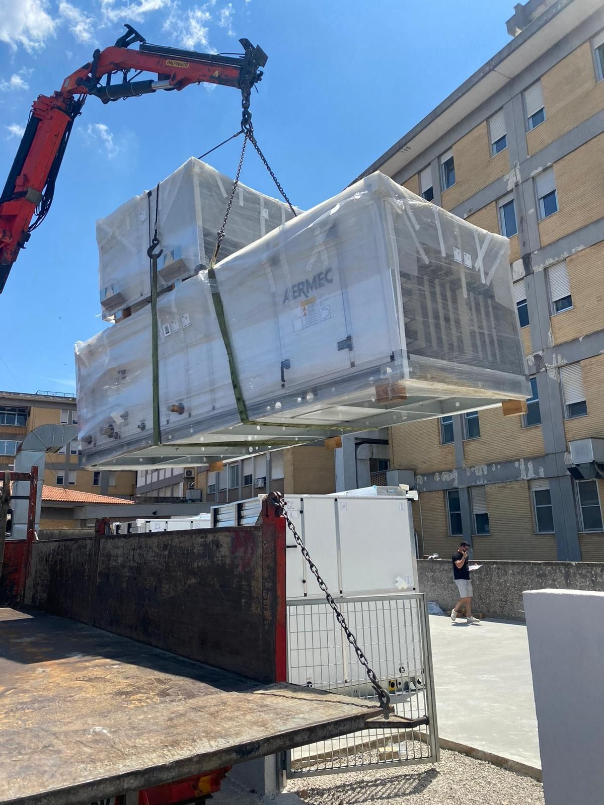 Una gru solleva un carico di pallet imballato accanto a un edificio in una strada soleggiata.