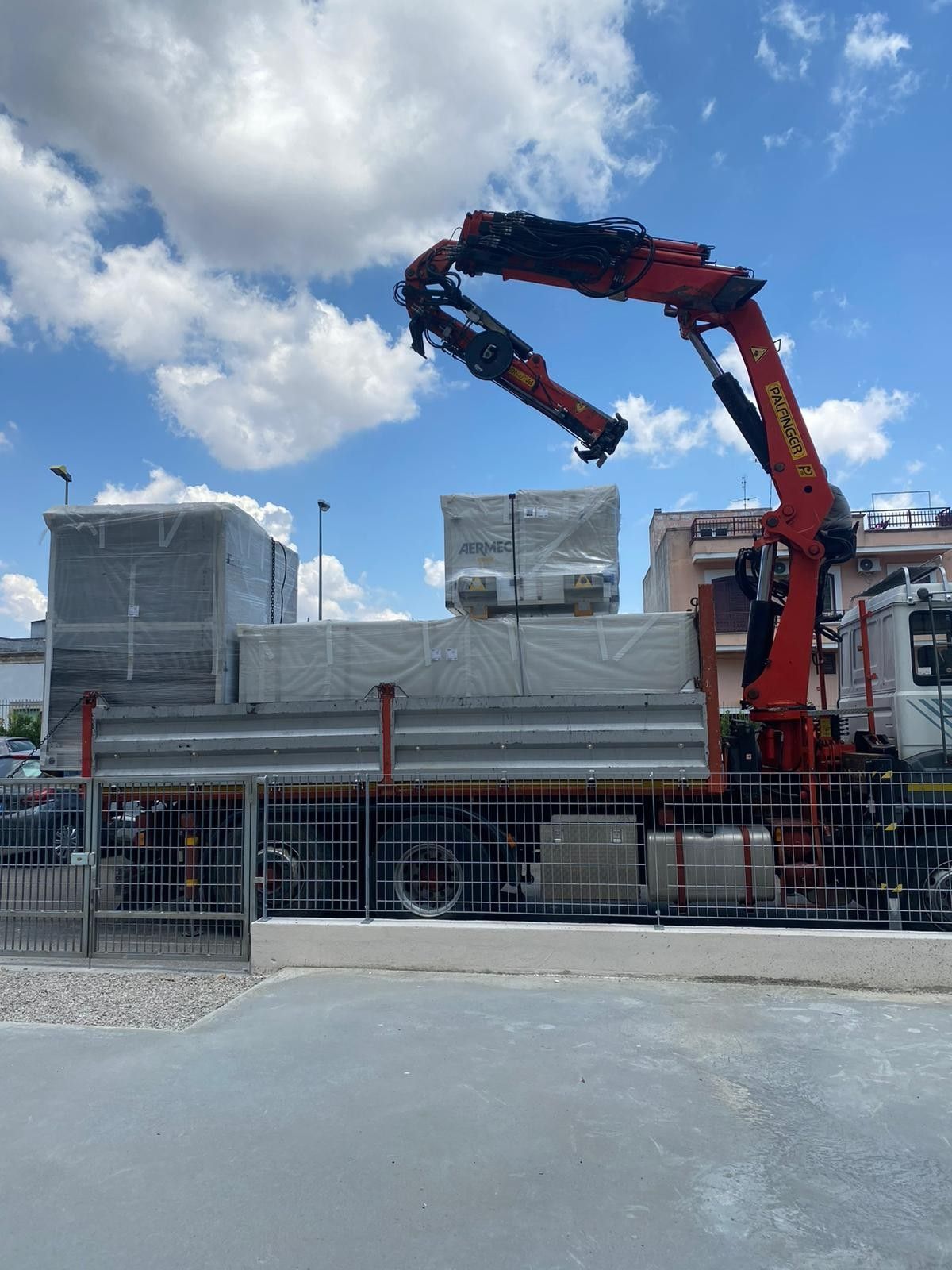 Una gru rossa solleva materiali edili imballati su un camion a pianale in un cantiere sotto un cielo azzurro nuvoloso.
