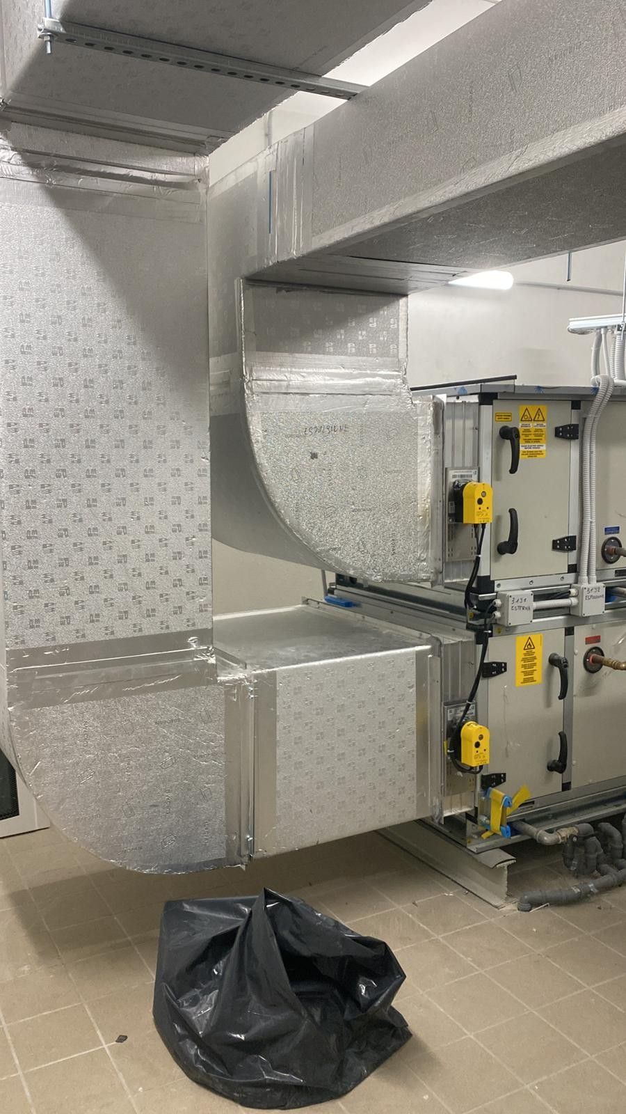 Condotti industriali e macchinari in un locale di servizio, con un sacco della spazzatura nero sul pavimento.