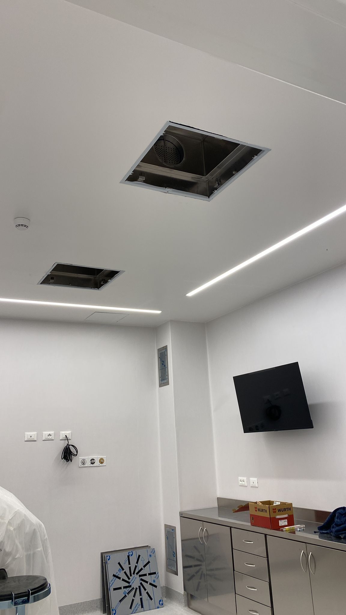 Camera moderna con pareti bianche, bocchette di ventilazione a soffitto, armadietti e TV a parete sulla destra.
