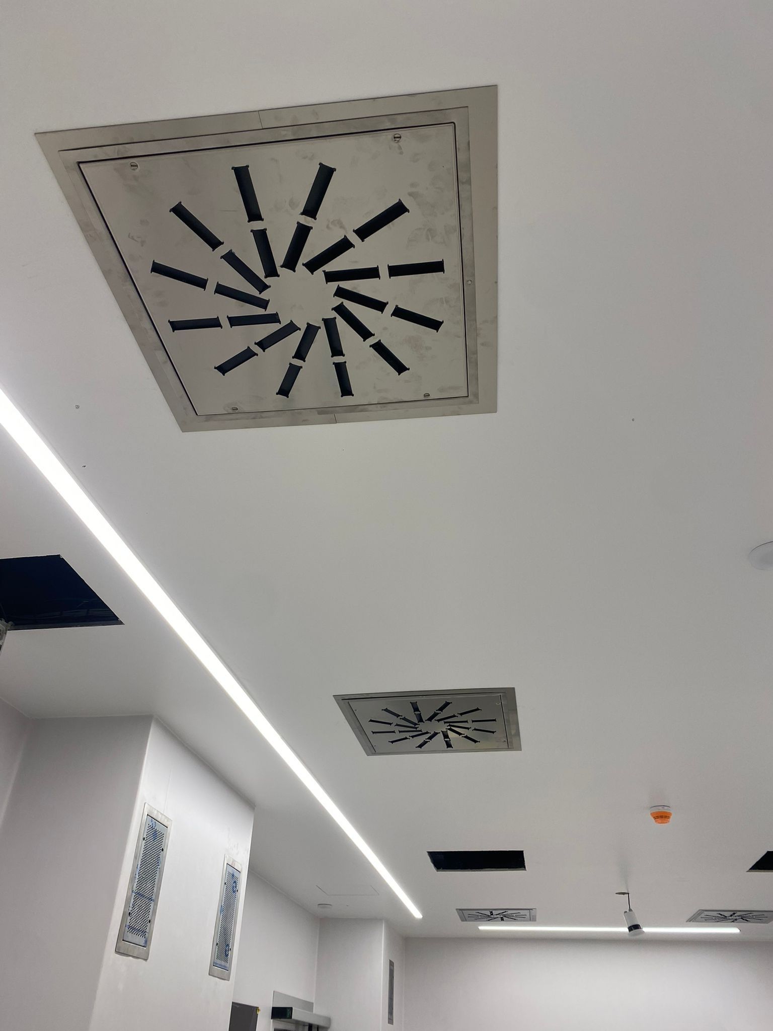 Soffitto bianco di un ufficio con bocchette di ventilazione quadrate e faretti a incasso, visto da un corridoio.