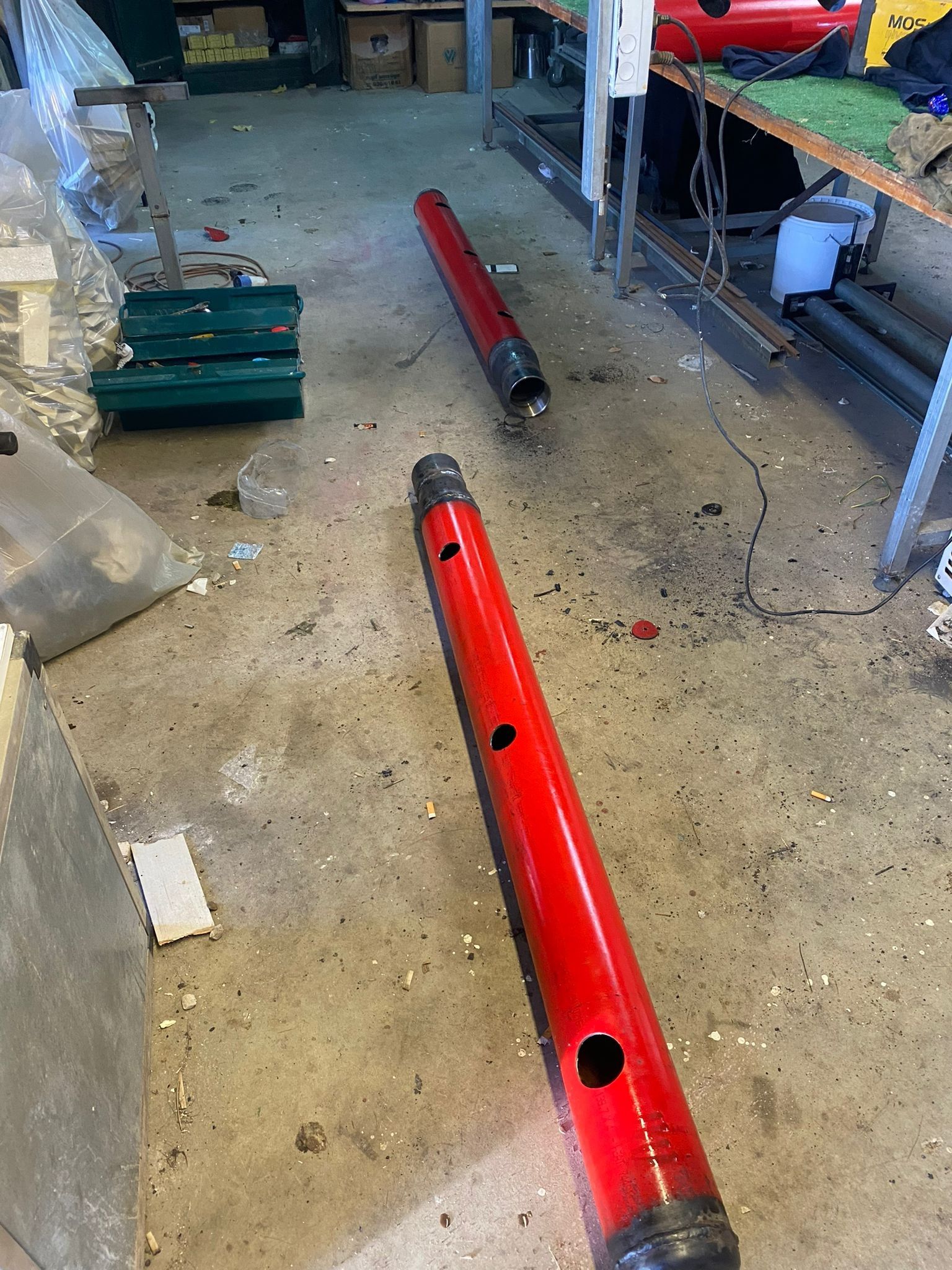 Pavimento dell'officina con due lunghi tubi metallici rossi, attrezzi e scaffali sullo sfondo.