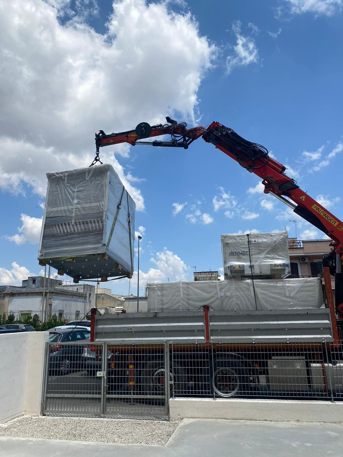 Una gru solleva un grande container metallico sopra un cantiere recintato sotto un cielo azzurro nuvoloso.