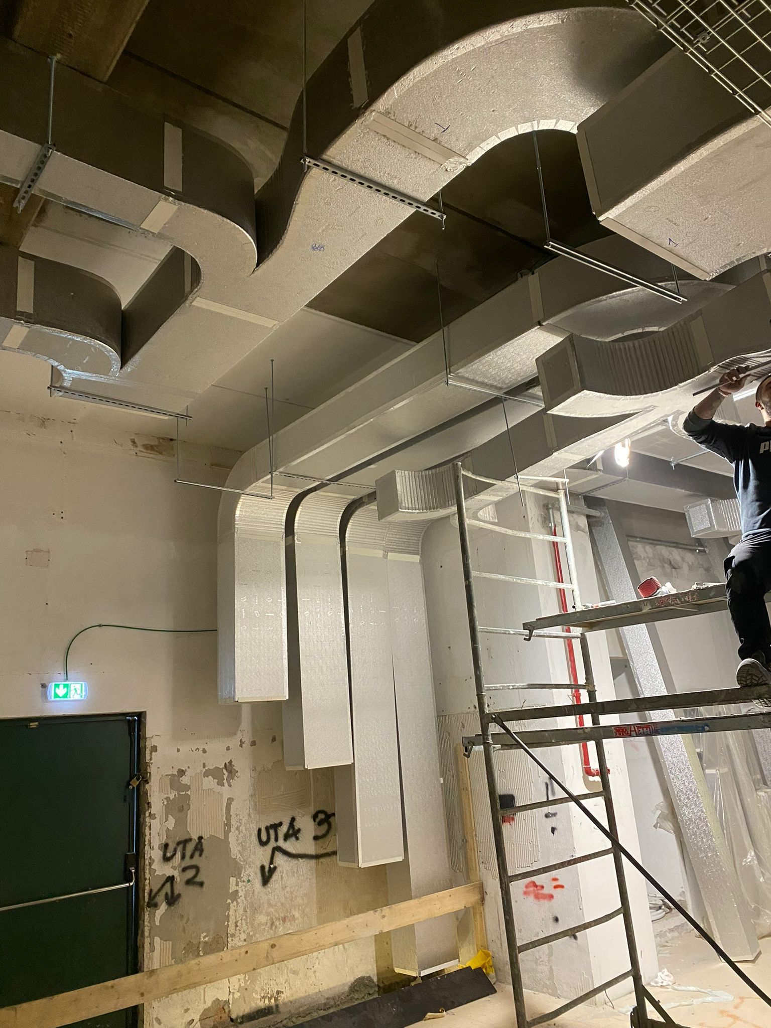 Cantiere edile interno con condotti a vista, scala e operaio su impalcatura vicino a un'apertura nel muro.