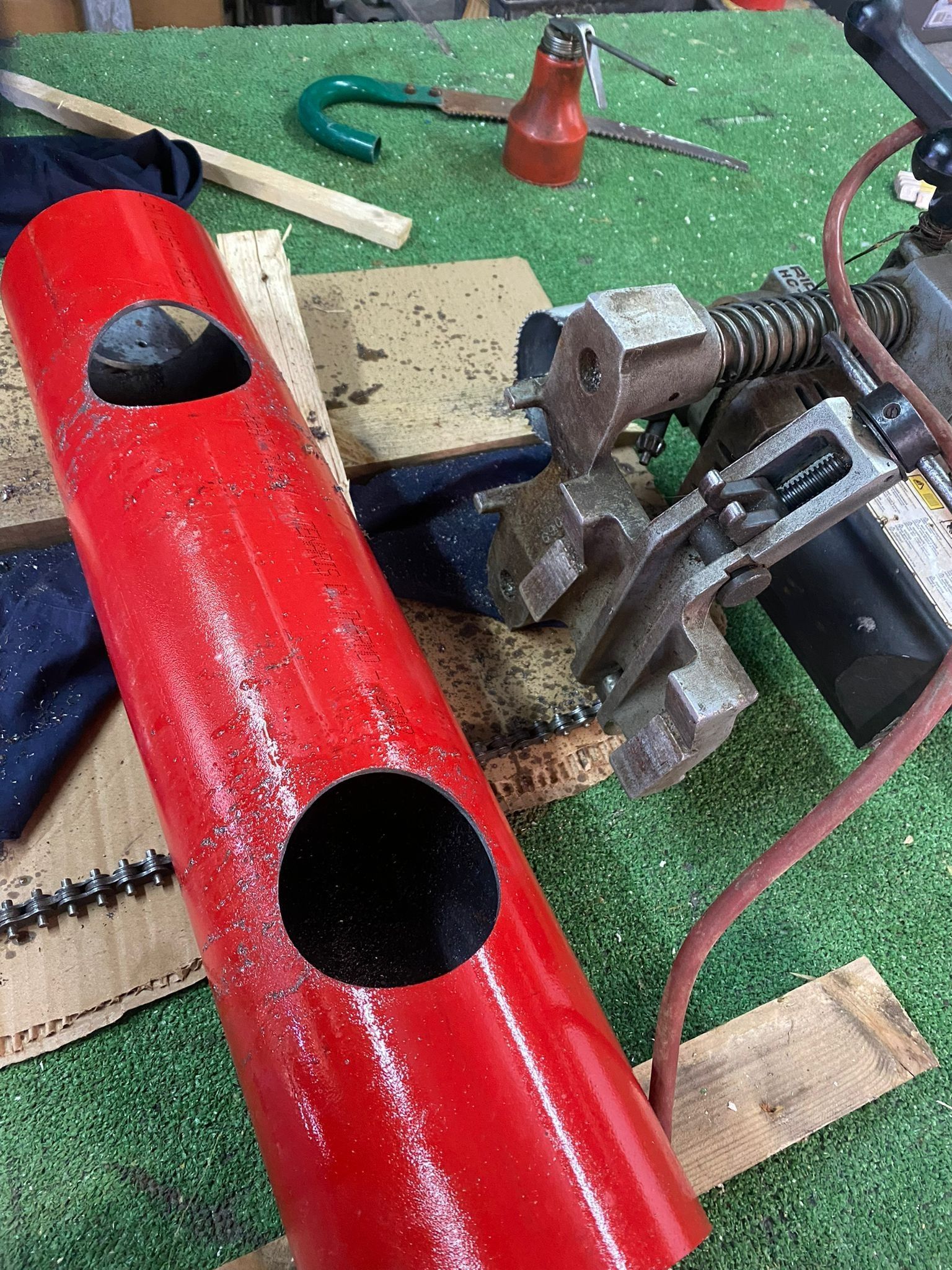 Tubo metallico rosso con intagli accanto a un morsetto su un banco da lavoro con segatura e attrezzi