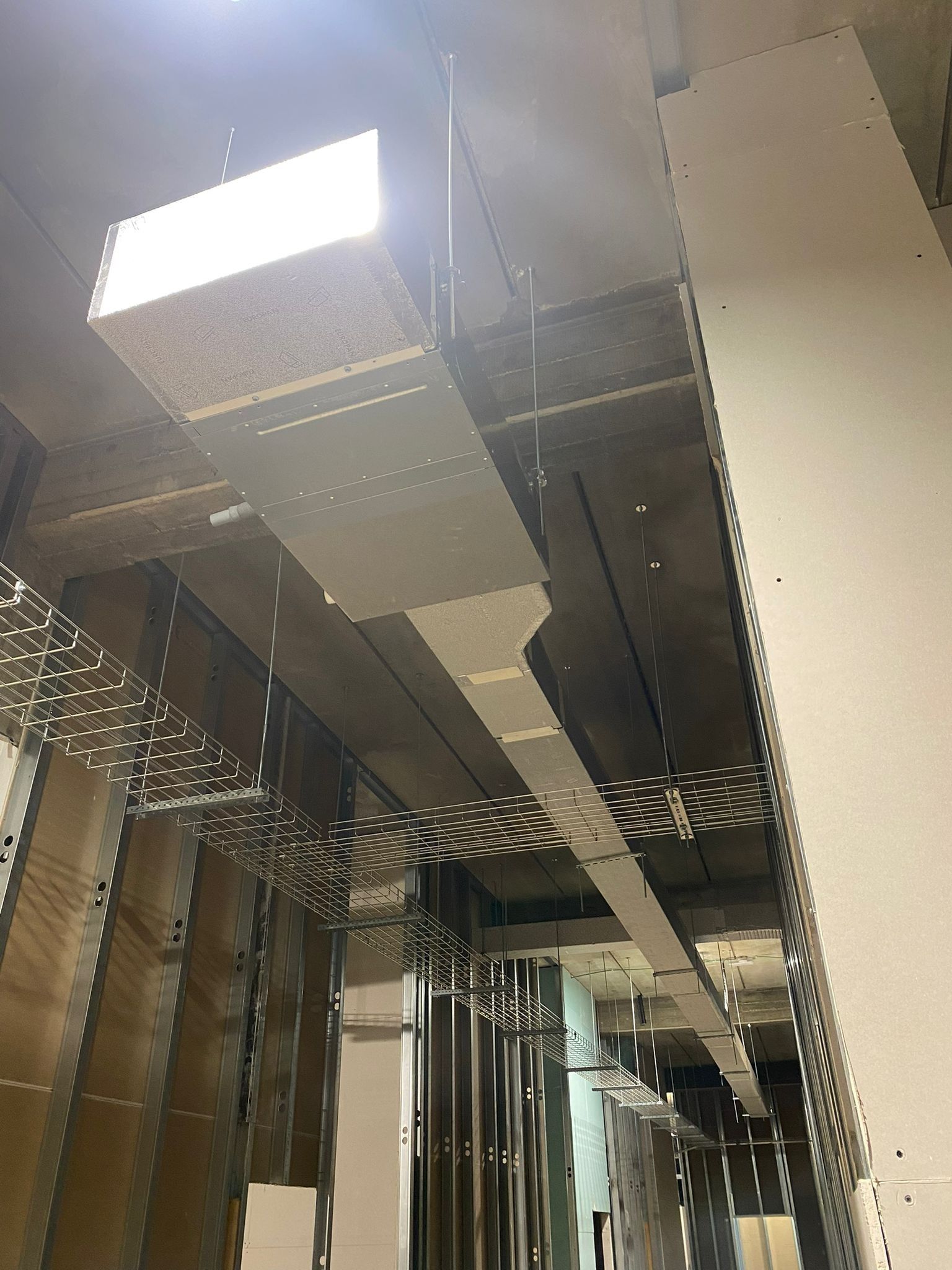 Vista dal soffitto di condotti di climatizzazione a vista e luci fluorescenti in un corridoio industriale.