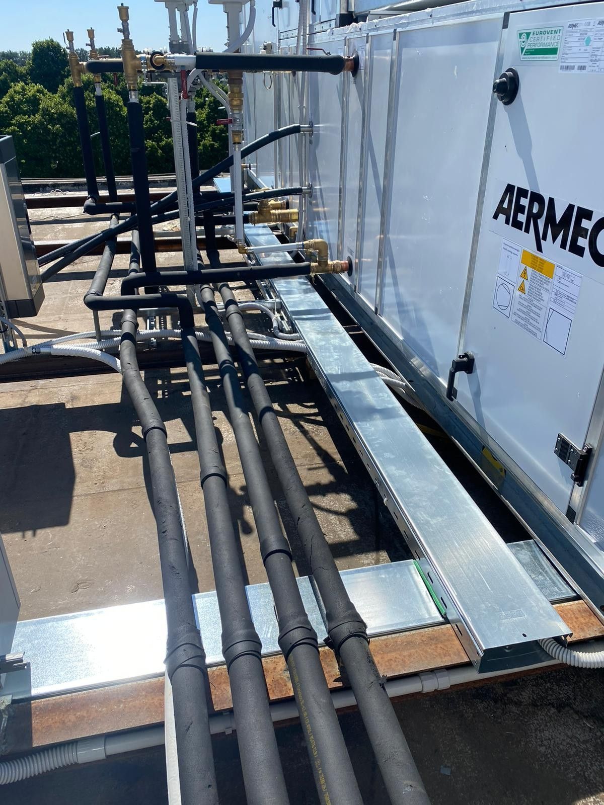 Tubature dell'impianto di climatizzazione e condotti metallici sul tetto, accanto a un'unità di trattamento aria bianca, in una giornata di sole.