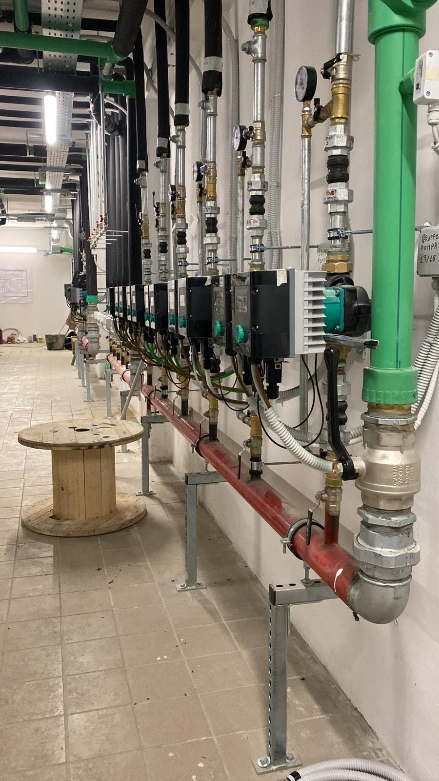 Tubazioni e valvole industriali lungo una parete bianca in un locale tecnico, con tubi verdi e una tubazione in cemento nelle vicinanze.