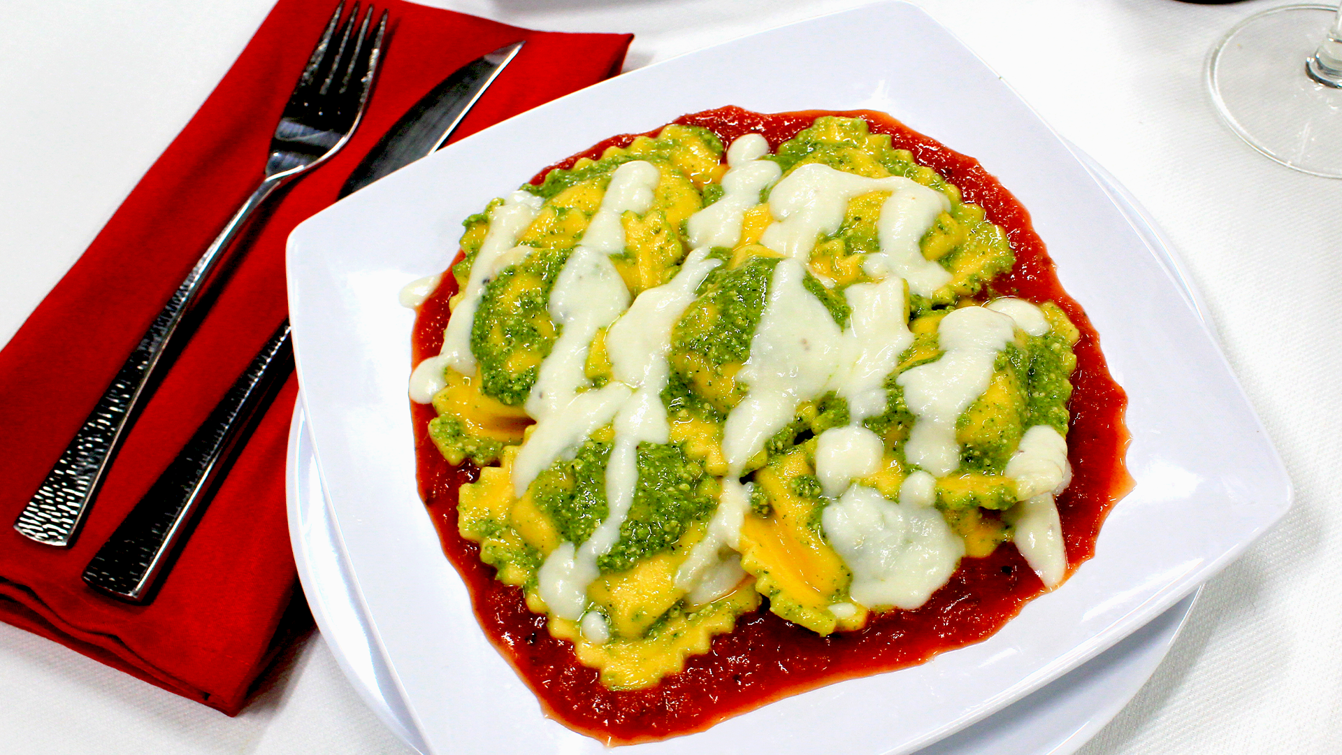 A plate of square ravioli topped with pesto and a creamy white sauce, served over a bed of red marinara sauce.