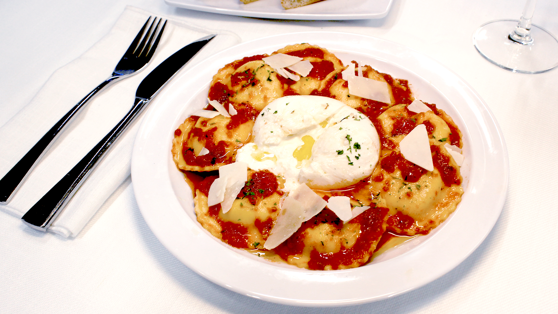 A plate of ravioli with tomato sauce, topped with burrata cheese and shaved parmesan, next to a fork and knife.