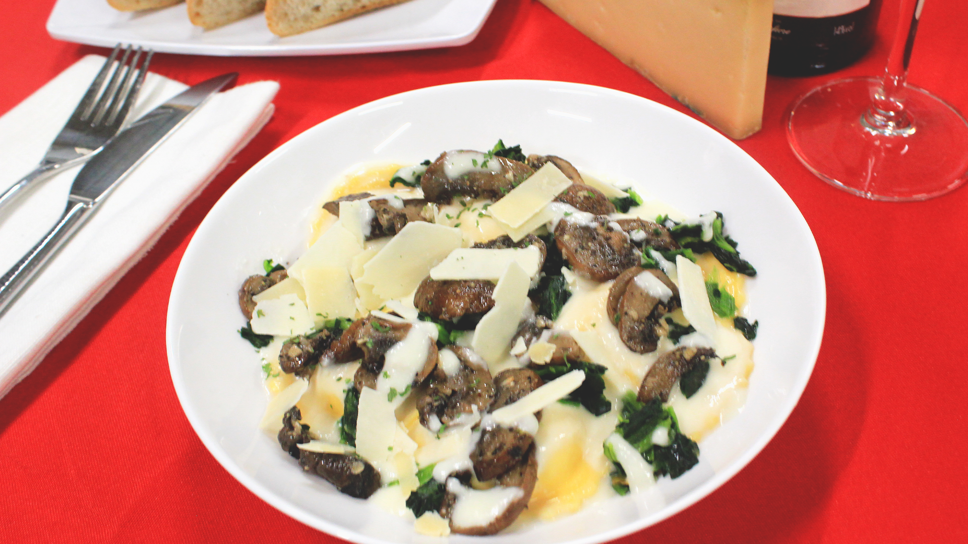 A white plate of sauteed mushrooms, greens, and shaved parmesan cheese sits on a bright red tablecloth.