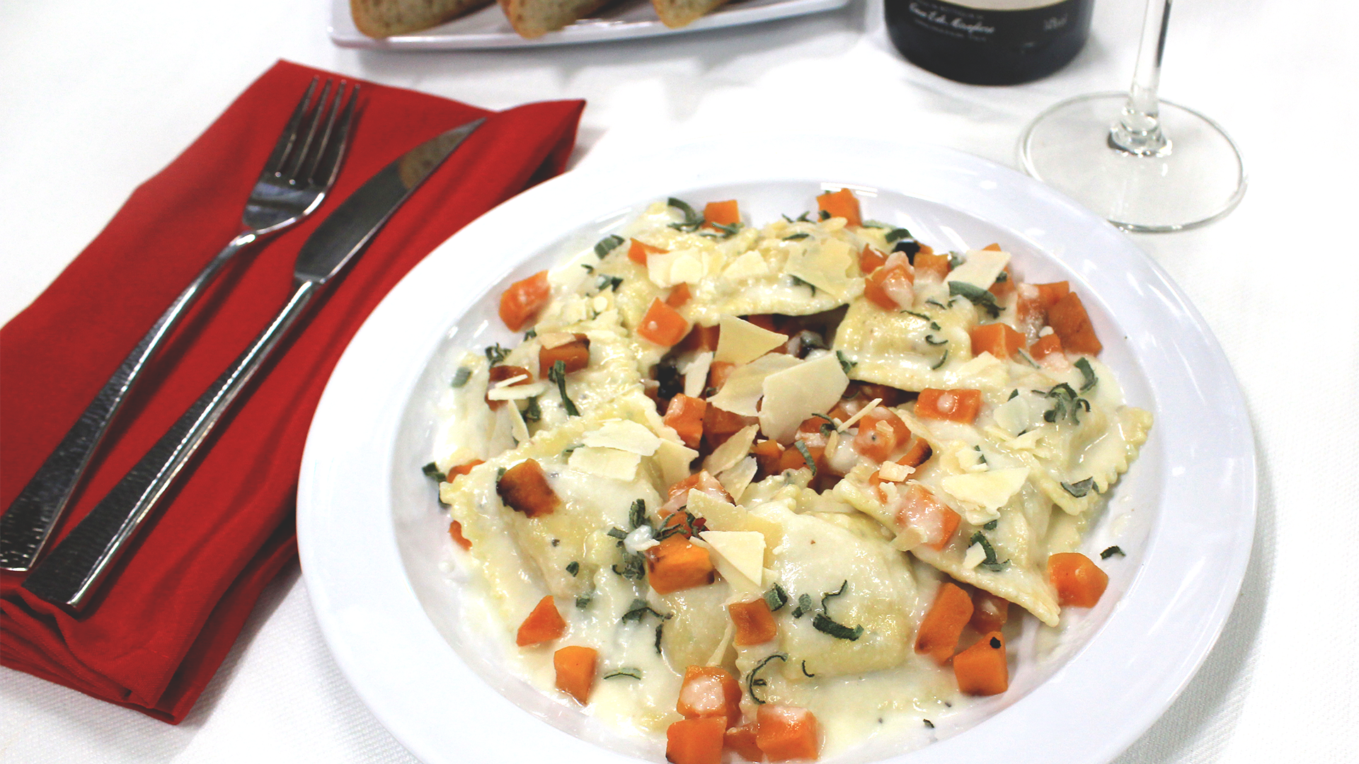 A plate of ravioli with a creamy white sauce, topped with diced carrots, herbs, and shaved cheese, beside red napkins.