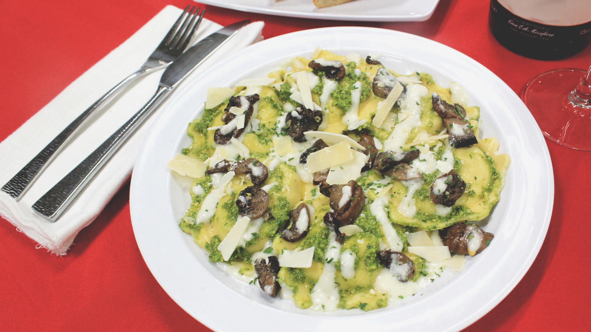 A white plate of pasta with pesto, mushrooms, and shaved cheese, set on a red tablecloth with a fork and knife.