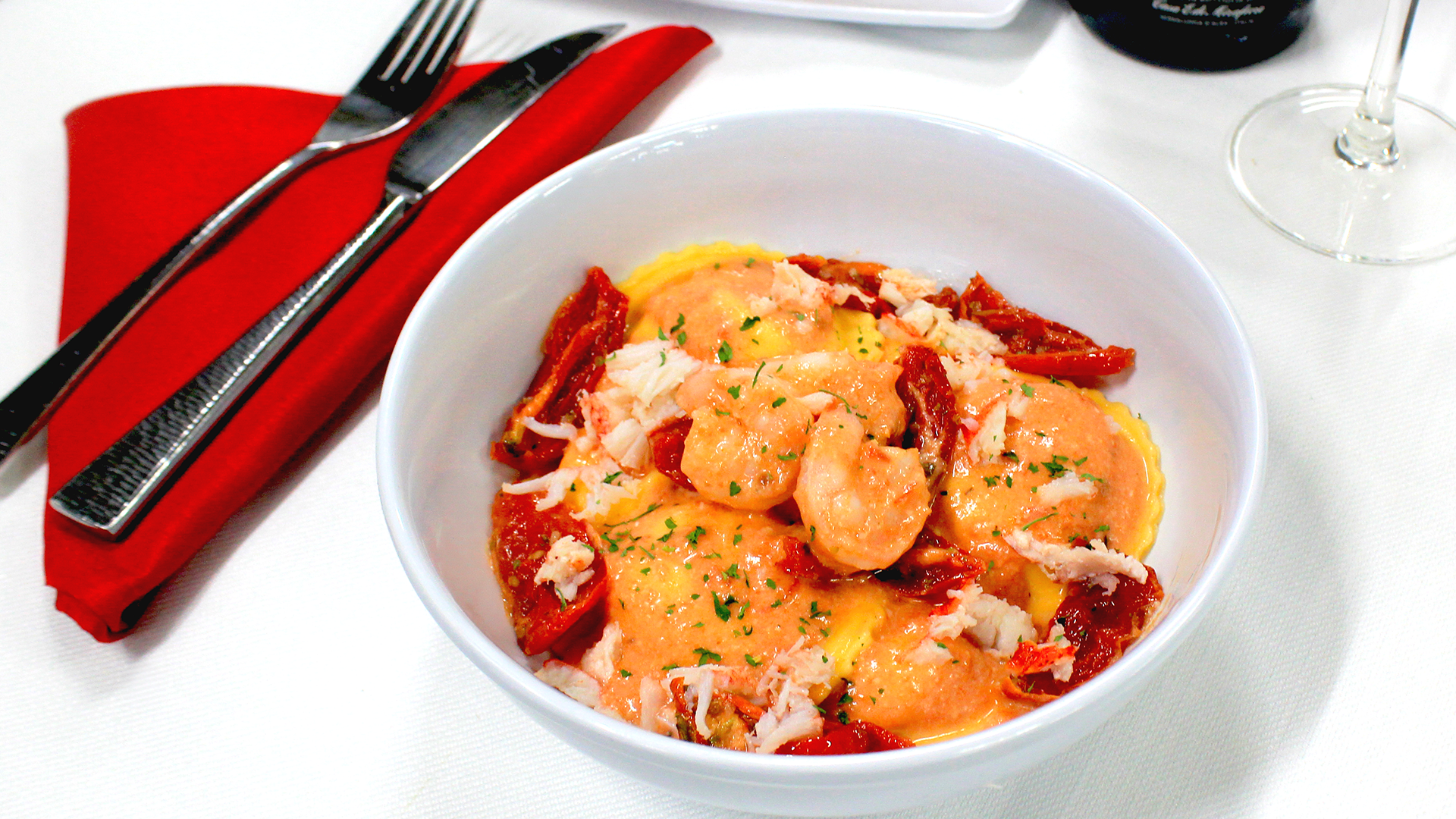 A white bowl of shrimp ravioli topped with crab and sun-dried tomatoes, set next to silverware on a red napkin.