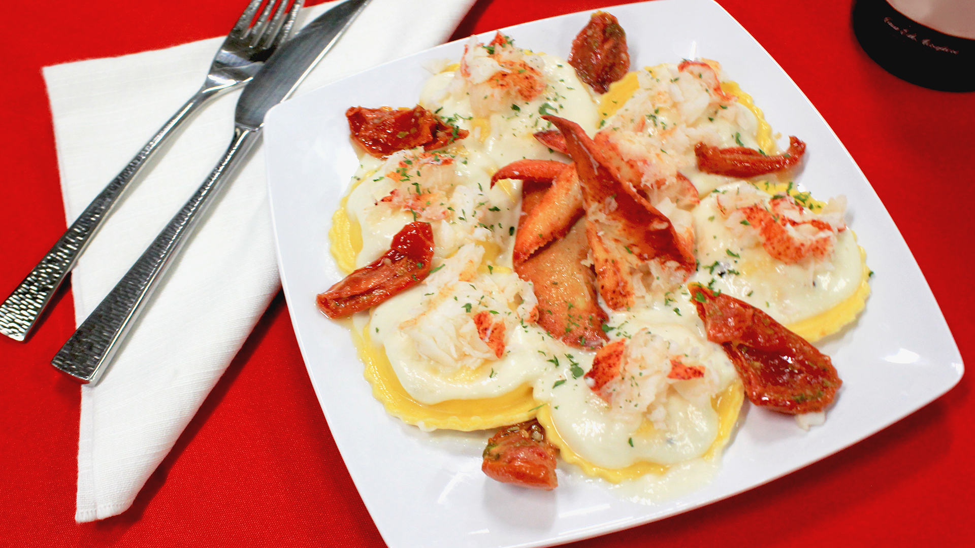 A plate of lobster ravioli in white sauce with sun-dried tomatoes, served with silverware on a vibrant red tablecloth.