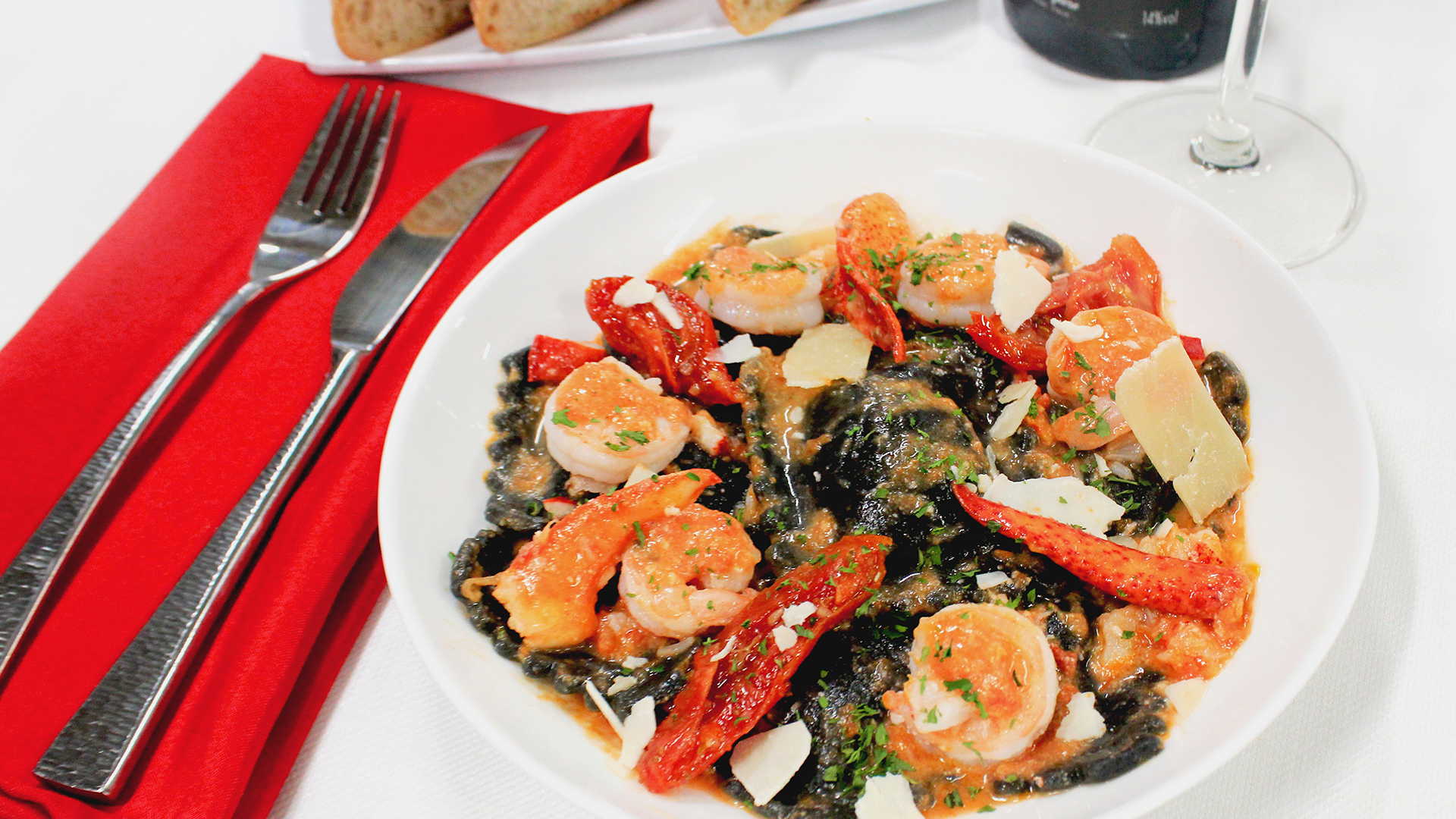 Black pasta with shrimp and sun-dried tomatoes on a white plate, served with a fork and knife on a red napkin.