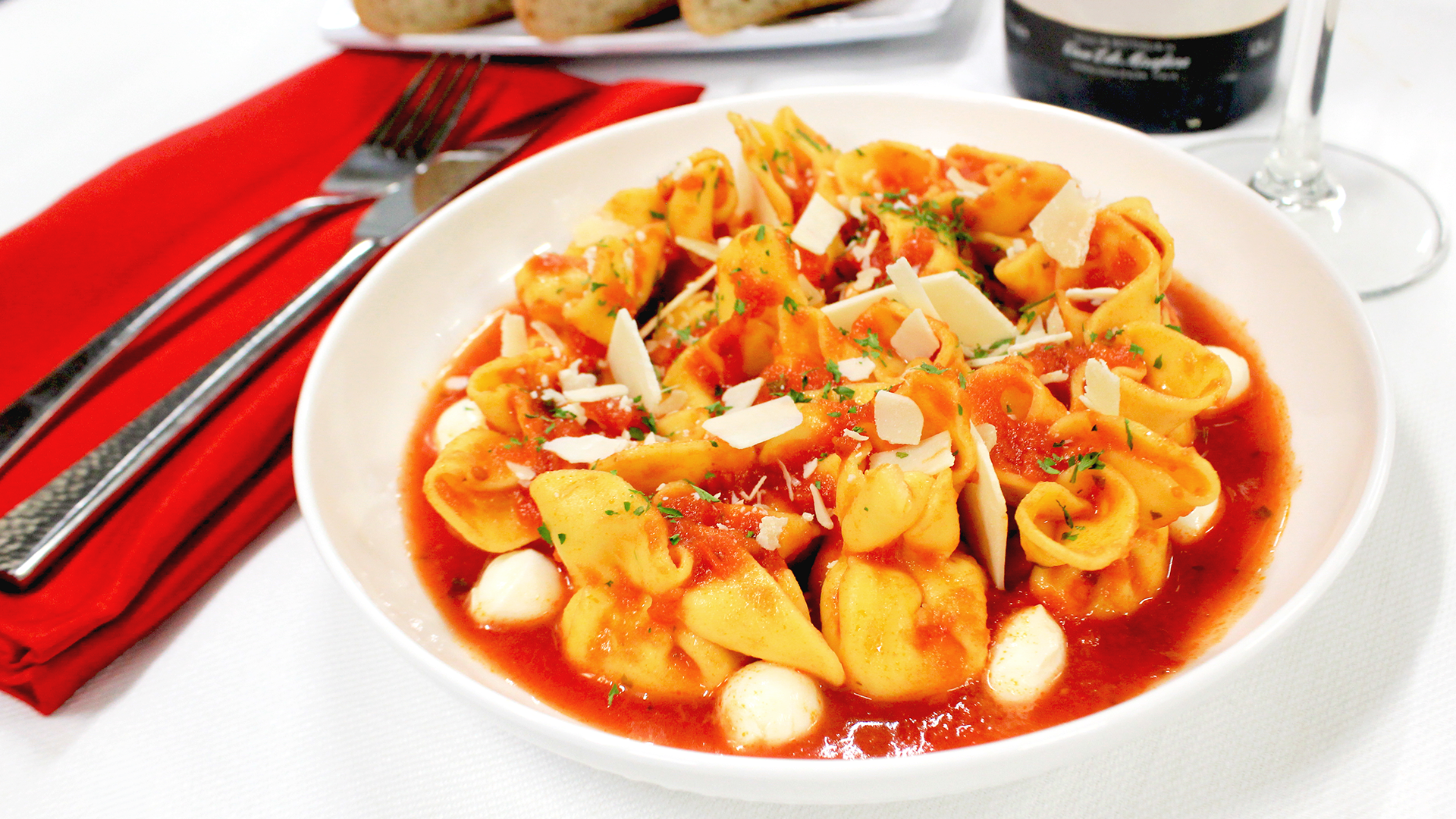 A bowl of tortellini pasta in red tomato sauce with mozzarella balls and parmesan shavings, beside a red napkin and fork.