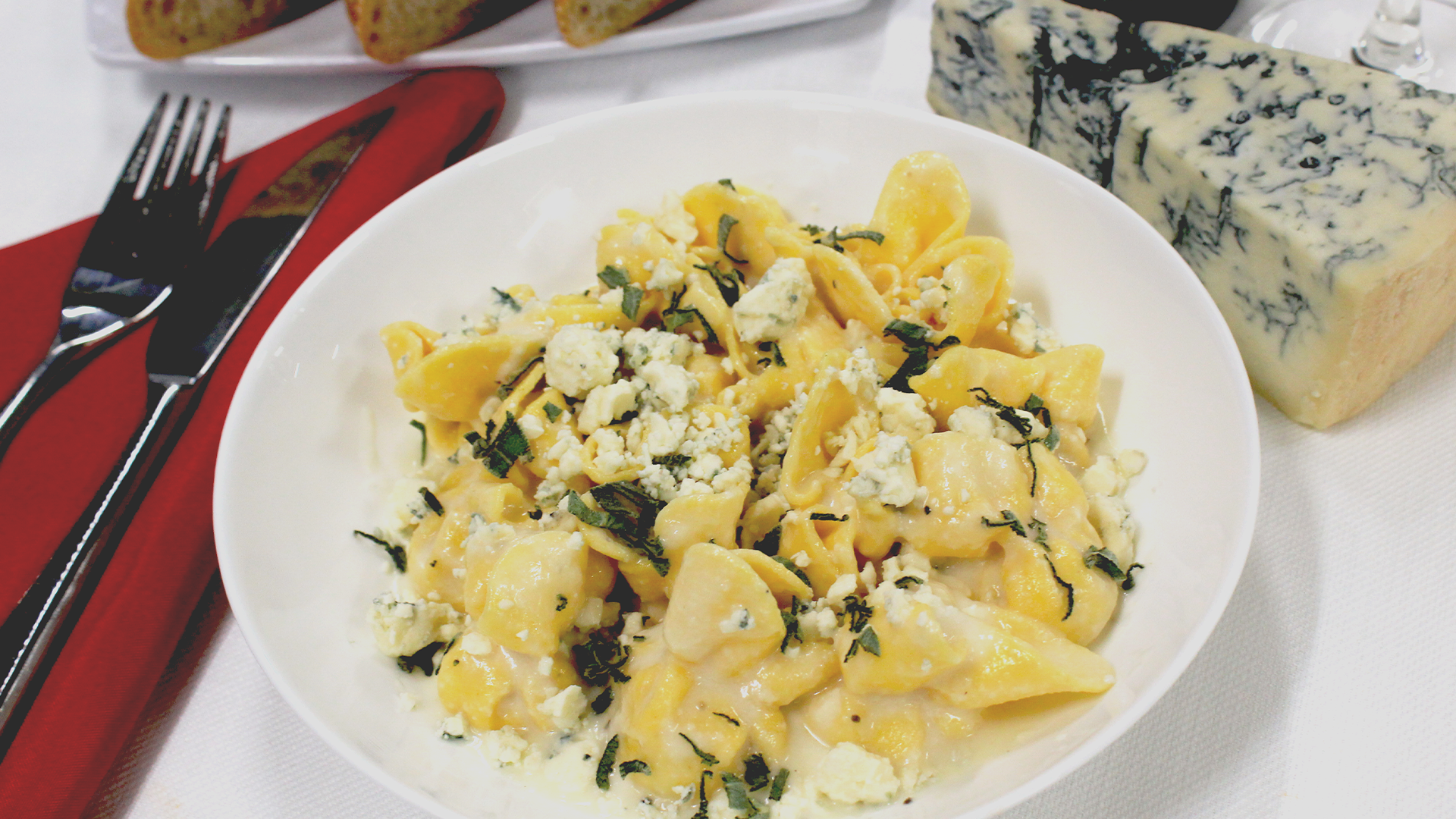 A bowl of pasta with blue cheese and herbs, next to a wedge of blue cheese, a red napkin, and a fork.