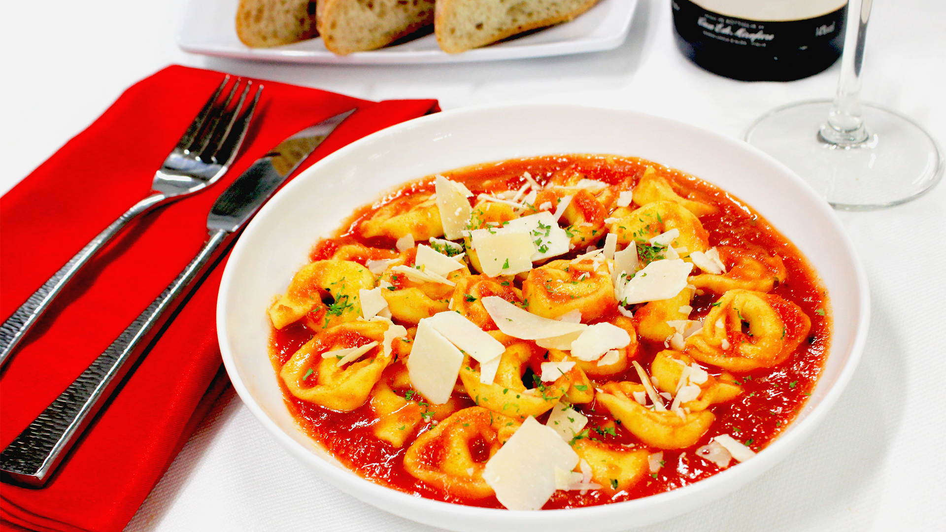 A bowl of cheese tortellini in tomato sauce, topped with parmesan shavings, served with bread and a glass of wine.