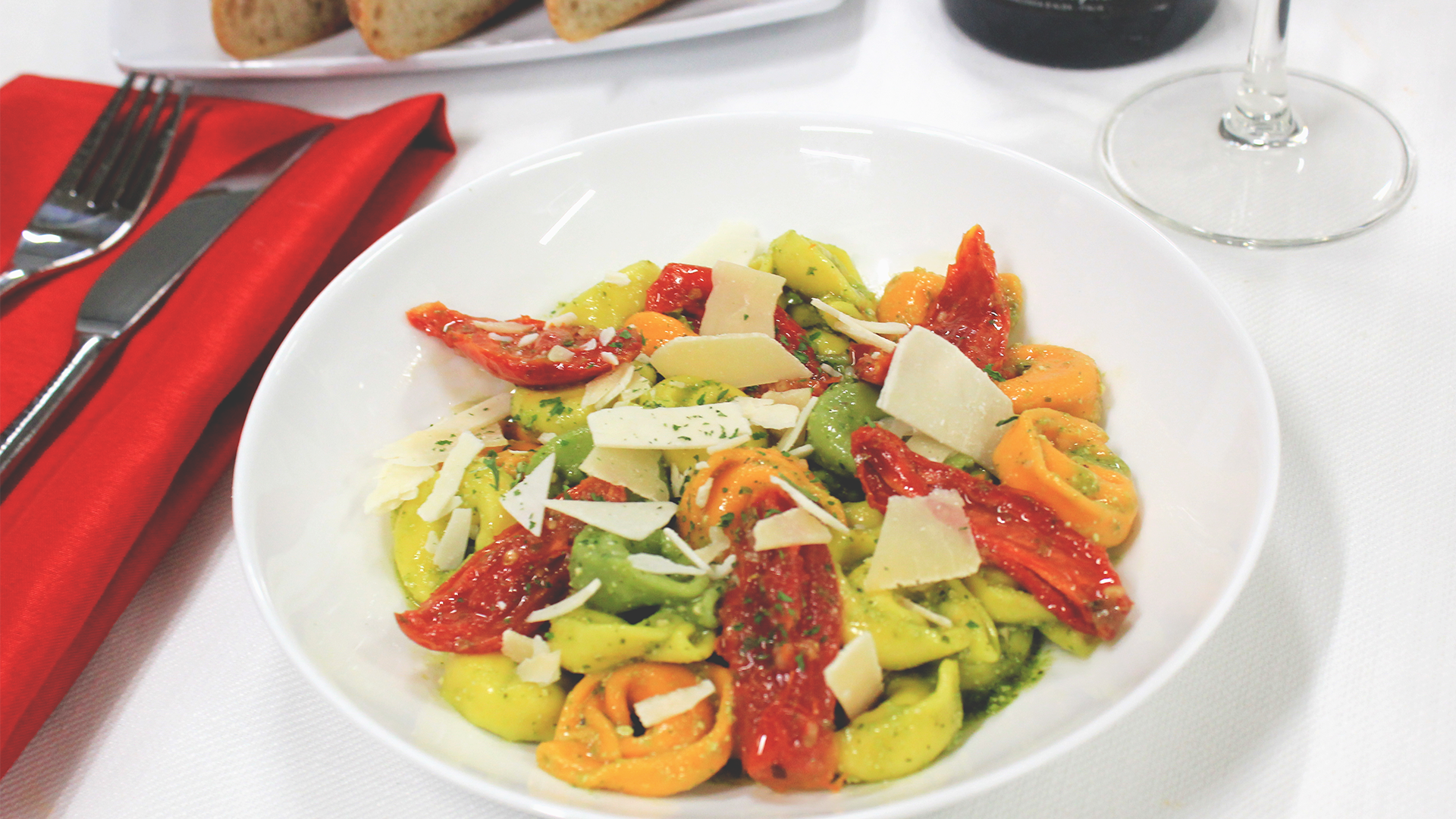 A bowl of tortellini with pesto, sun-dried tomatoes, and parmesan shavings sits next to cutlery and a red napkin.