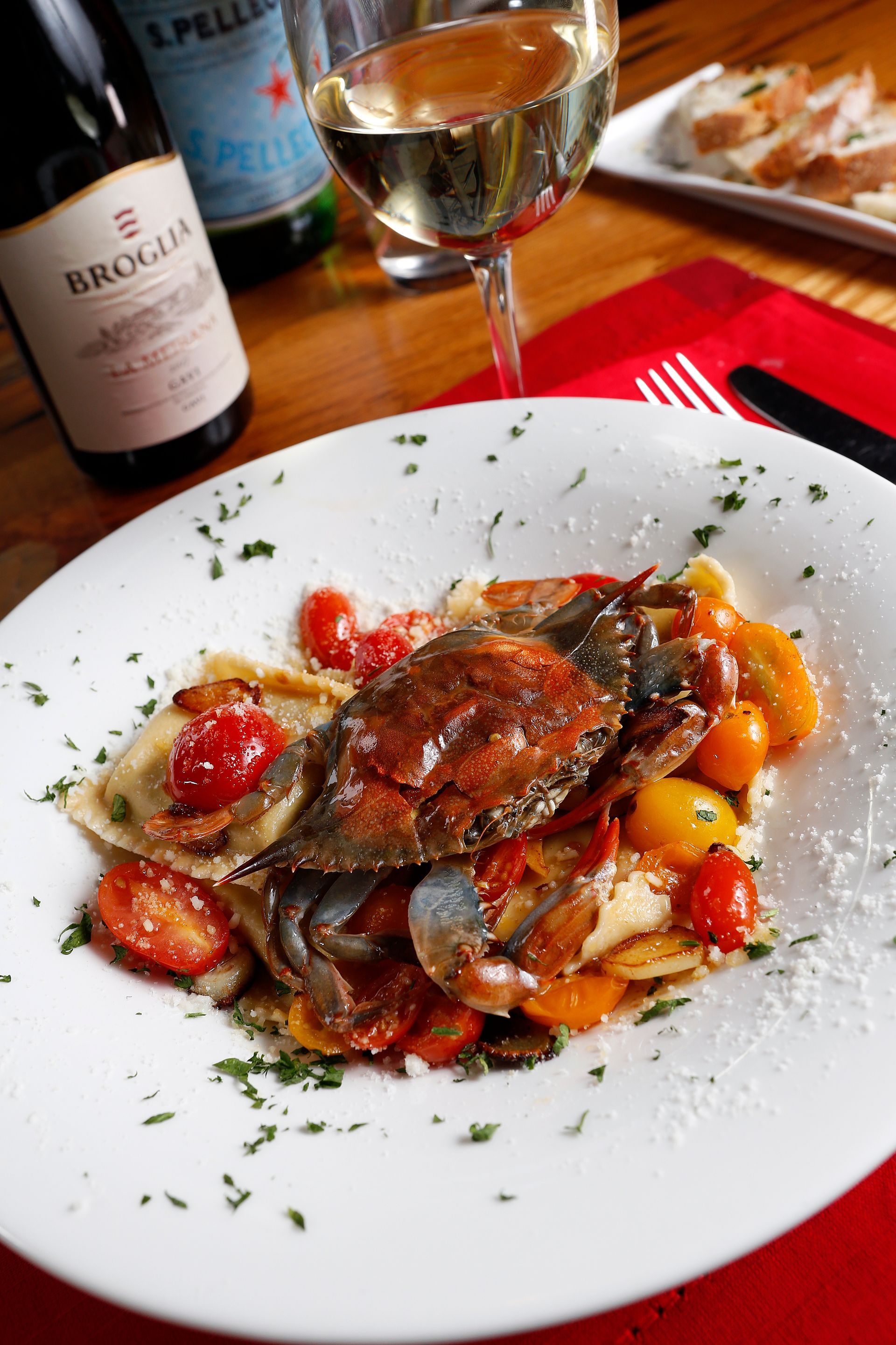 A close-up of a soft-shell crab dish with cherry tomatoes and pasta on a white plate, served with wine and bread.