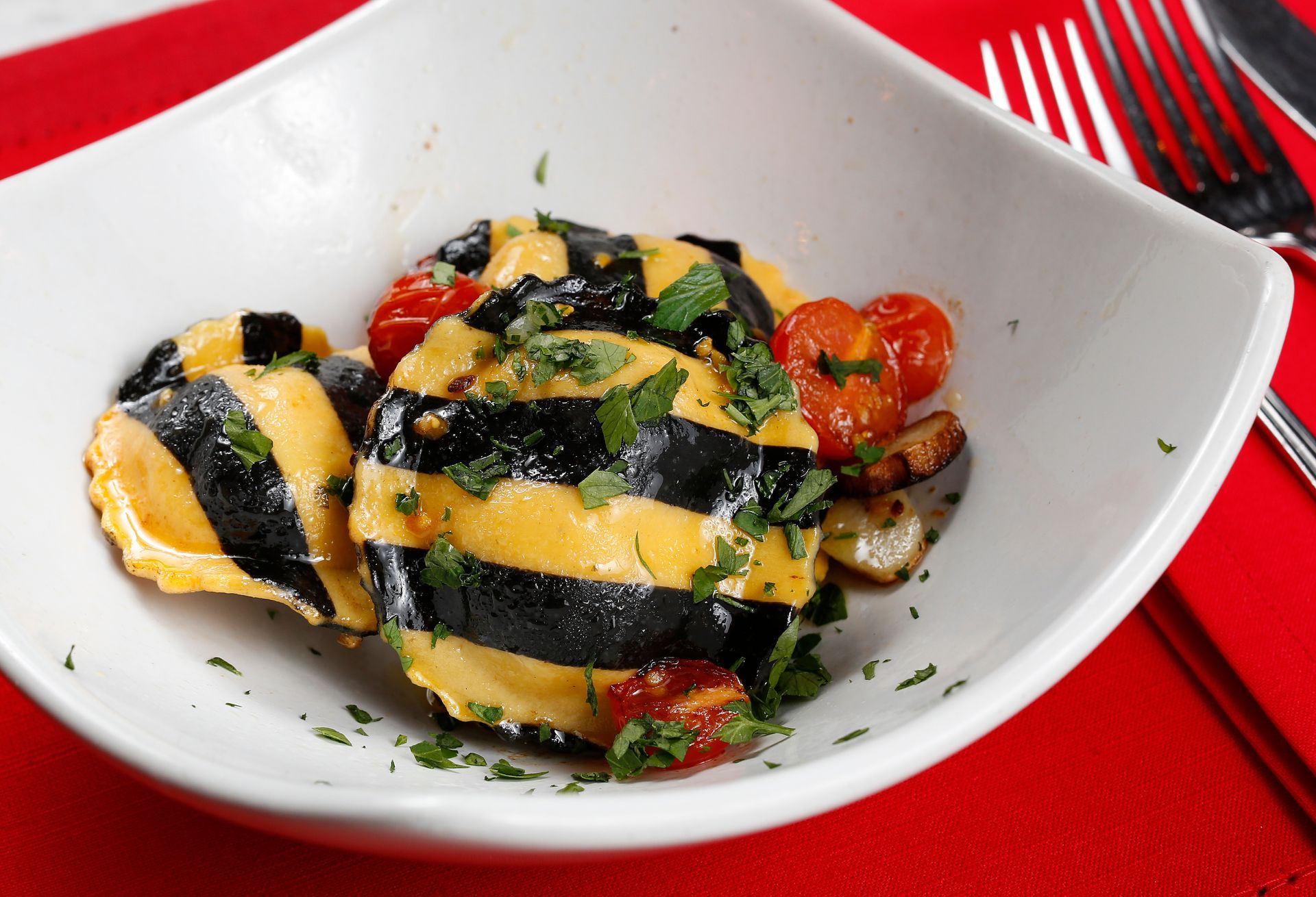 Striped ravioli with roasted cherry tomatoes and fresh herbs in a white bowl on a bright red background.