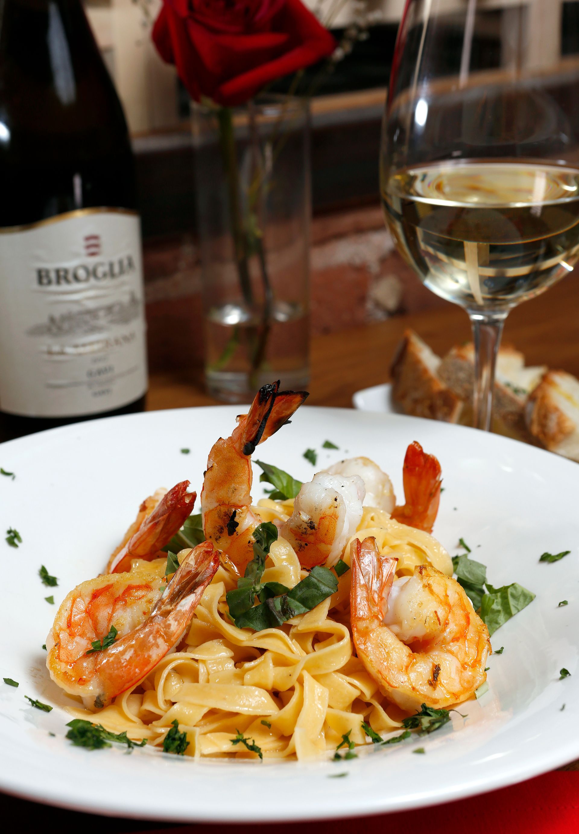 Shrimp scampi pasta served in a white bowl, with a wine bottle, a glass of white wine, and a red rose in the background.