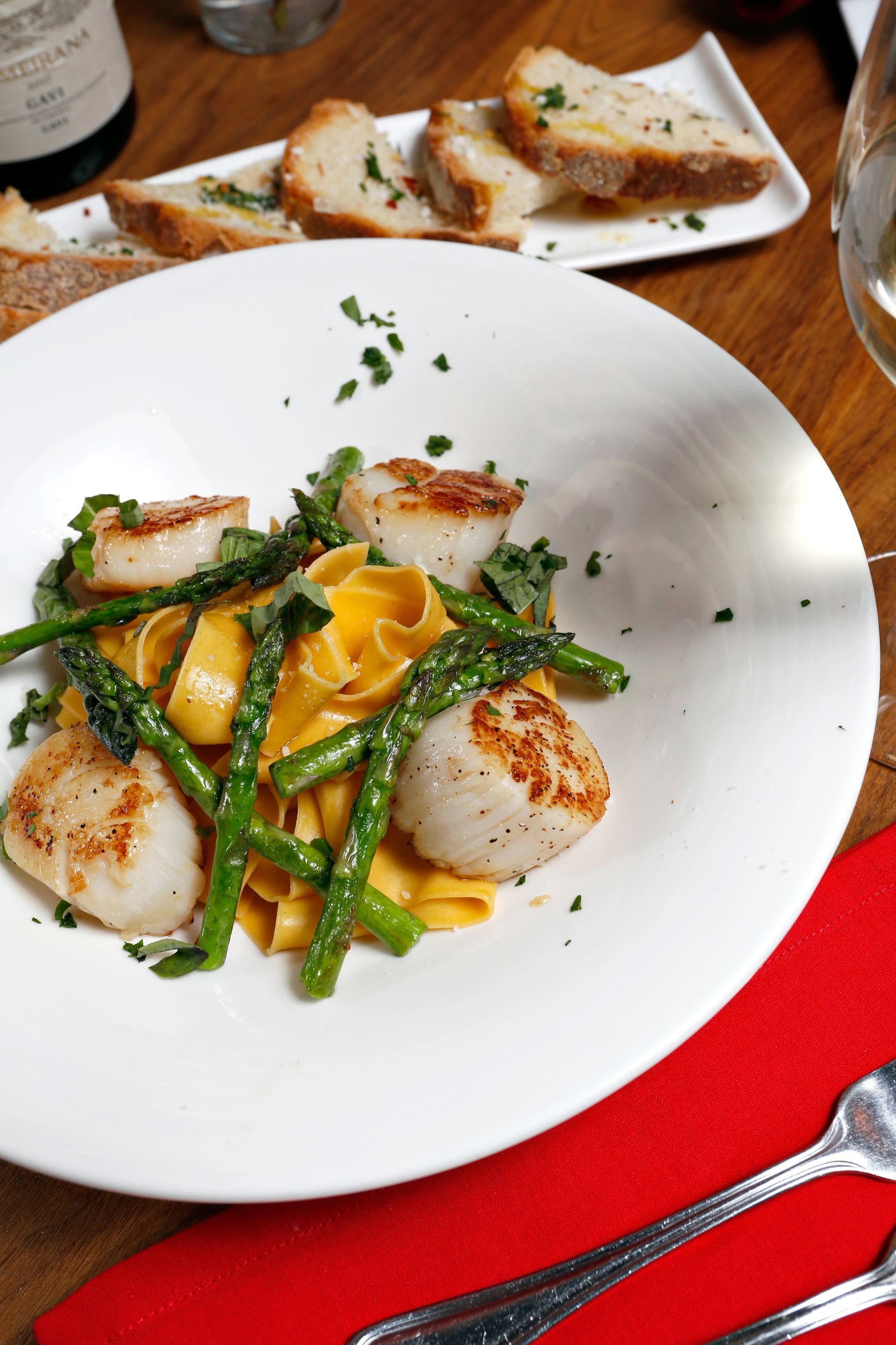 A dish of seared scallops and asparagus over yellow pasta in a white bowl, with bread on a side plate.
