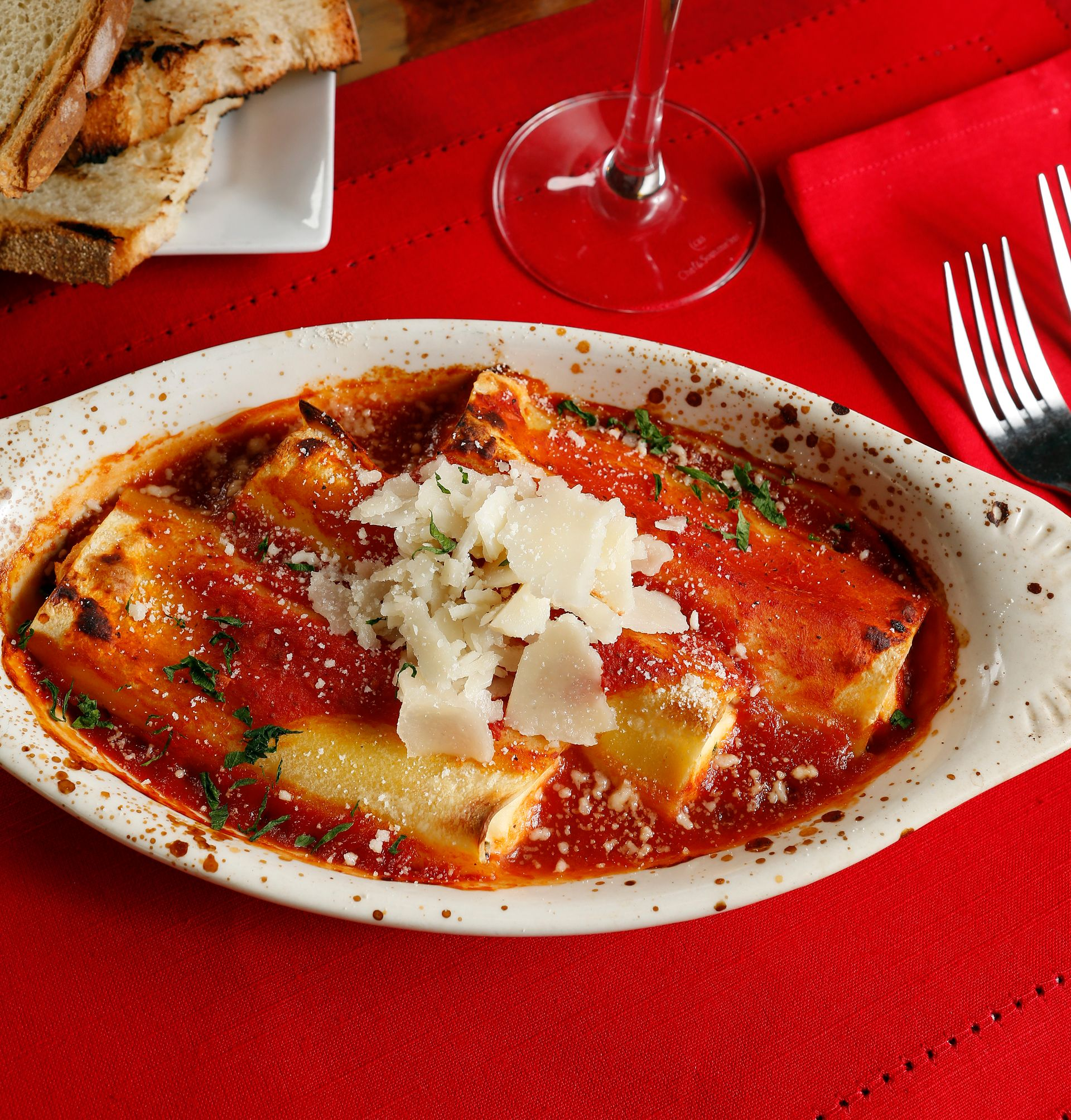 A serving of baked pasta tubes in red tomato sauce, topped with shaved cheese, on a table with bread and a red napkin.