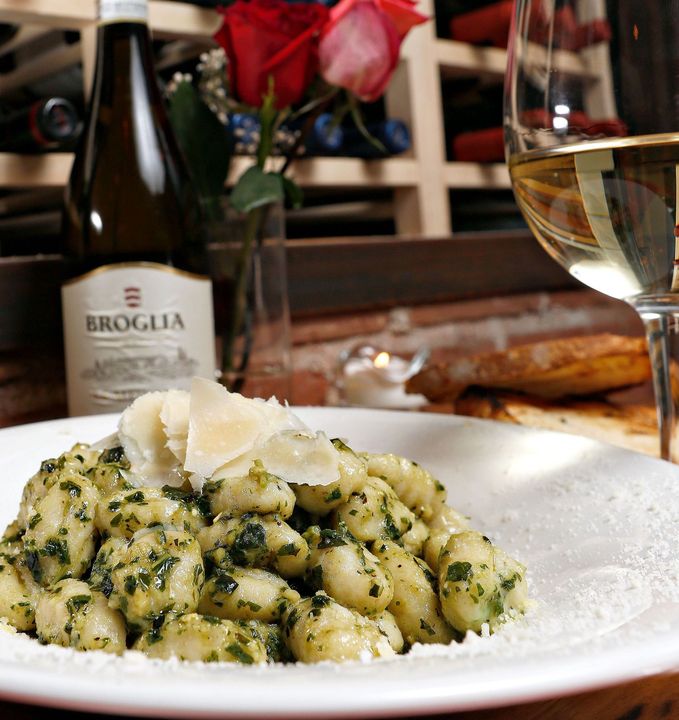 A plate of gnocchi with pesto and shaved cheese next to a glass and bottle of wine, with roses in the background.