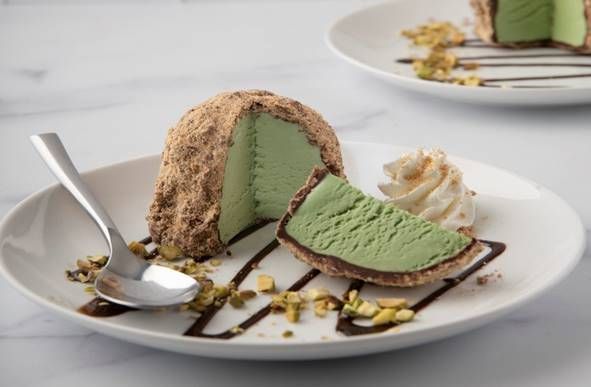 A scoop of light green pistachio ice cream coated in crumbs, sliced open on a white plate with chocolate drizzle and nuts.