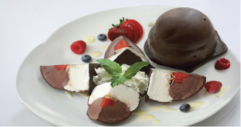 A plated dessert with a whole chocolate-covered dome alongside slices filled with white cream, berries, and mint.