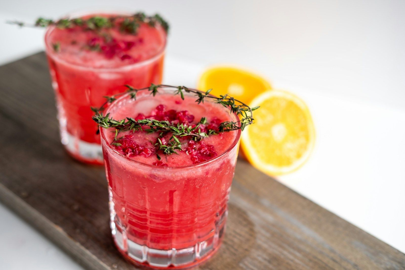 Two glasses of bright red fruit cocktail garnished with sprigs of thyme, placed on a wooden board with citrus slices.