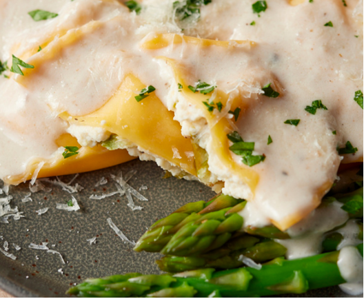 A plate of ravioli topped with creamy white sauce and parsley, served alongside fresh green asparagus spears.