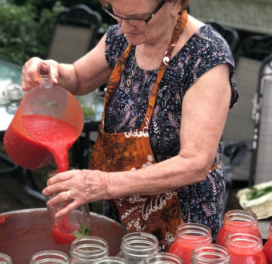 A person wearing an apron pours bright red tomato sauce from a pitcher into a glass jar among several others.