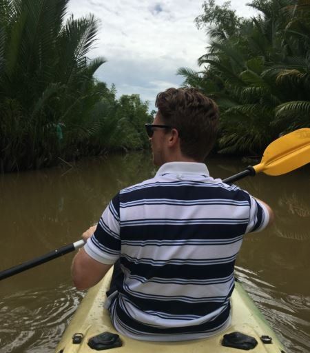 Een persoon, gekleed in een gestreept poloshirt en een zonnebril, peddelt in een kajak door een smalle, weelderige waterweg omzoomd met palmbomen.