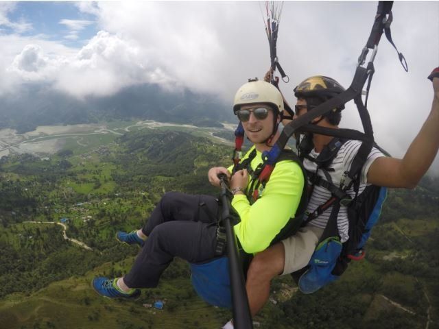 Twee mensen maken een tandemparaglidingvlucht boven een groen, bergachtig landschap met wolken aan de hemel.